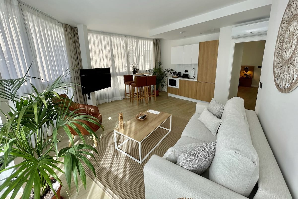 Una sala de estar moderna con sofá, mesa de centro y televisor. Cuenta con una zona de cocina con armarios de madera y una mesa de comedor.