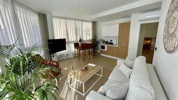 Una sala de estar moderna con sofá, mesa de centro y televisor. Cuenta con una zona de cocina con armarios de madera y una mesa de comedor.