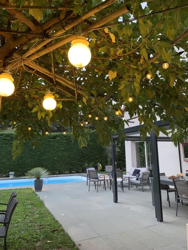 An outdoor patio area with a pergola, string lights, seating, and a pool.