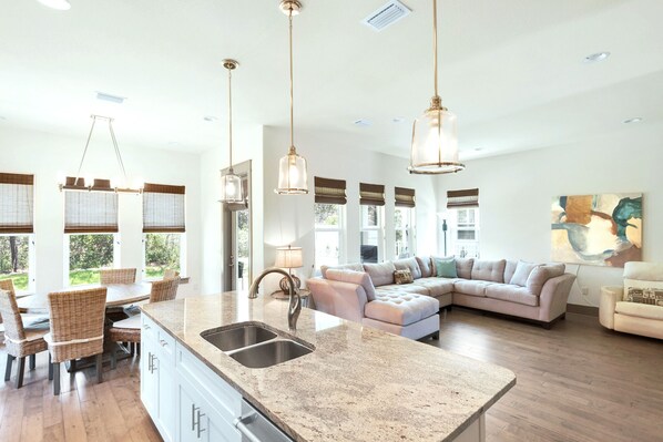 A spacious kitchen with a large island, a dining area with wicker chairs, and a living room with a sectional sofa.