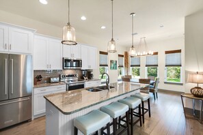 A modern kitchen with white cabinets, stainless steel appliances, a granite island with bar stools, pendant lighting, and large windows with views of greenery.