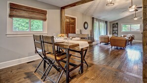 A dining area with a wooden table and chairs, a kitchen in the background, and a living room with sofas.