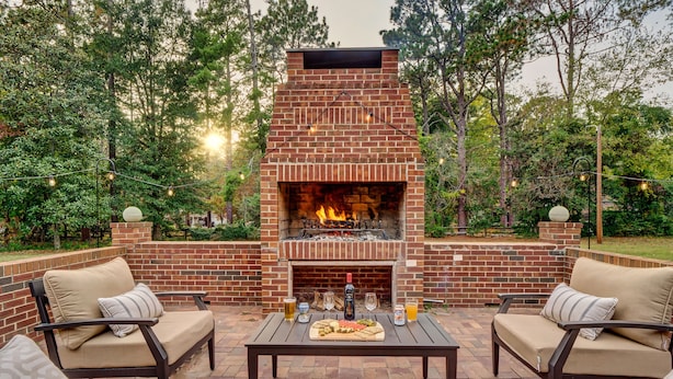An outdoor patio with a brick fireplace, two cushioned chairs, a table with food and drinks, surrounded by trees.