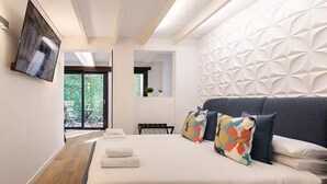 A modern bedroom with a large bed, a TV mounted on the wall, and a view of greenery outside. - Diagonal Amazing 3BD with private garden (Barcelona)