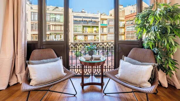A balcony with wicker chairs, a small table with a bowl and a plant, and a view of multi-story buildings. - Elegant and historic 3BD in Pl Urquinaona! (Barcelona)