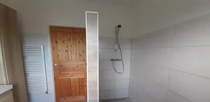 Une salle de bain avec une porte en bois, une douche équipée d’un pommeau de douche amovible et des murs recouverts de carreaux blancs.