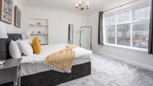 A bedroom with a bed, bedside tables, a nightstand, a chandelier, a window, and a shelf with decorative items.