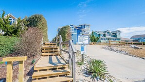 Een pand aan het strand met houten trappen naar een zandgebied, een bord met strandregels, en een uitzicht op blauwe huizen op de achtergrond.