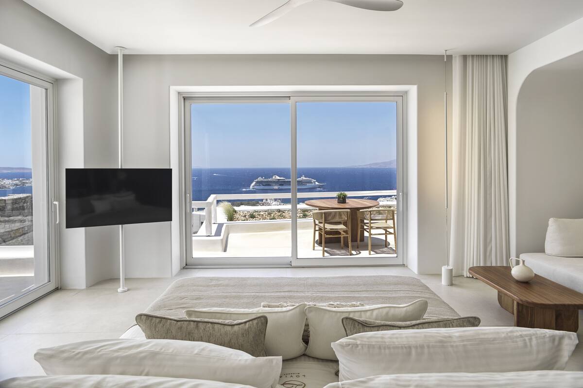 A modern hotel room with a large balcony, a flat-screen TV, and a cozy seating area with cushions.