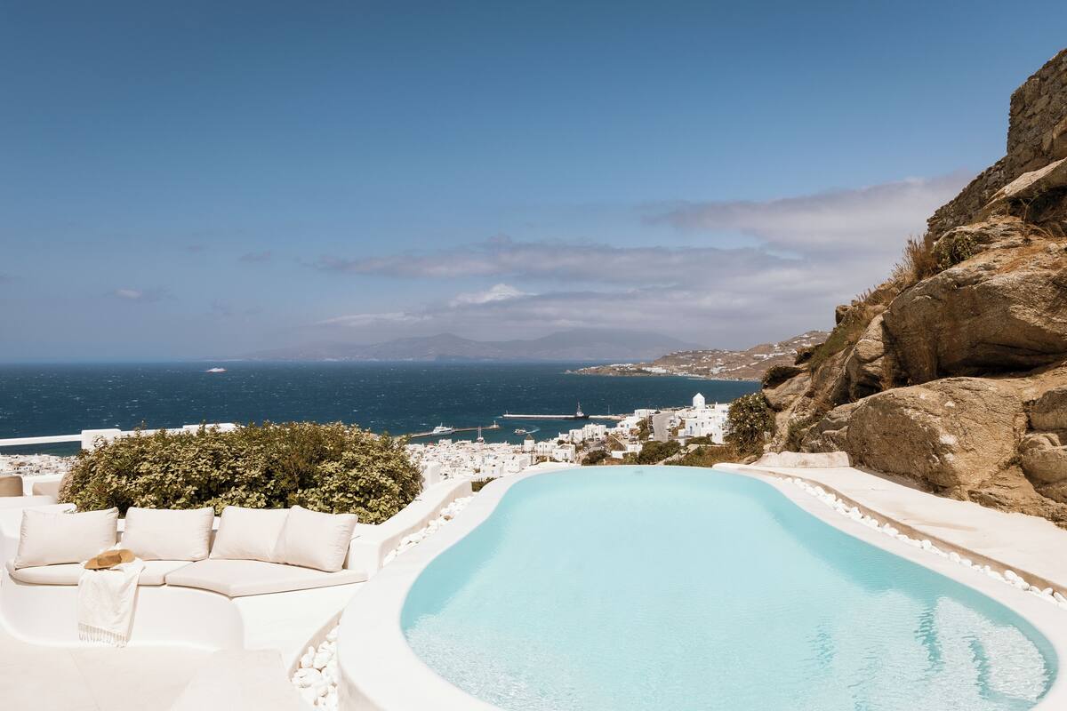A pool with a curved edge, white outdoor furniture, and a view of the sea and distant buildings.