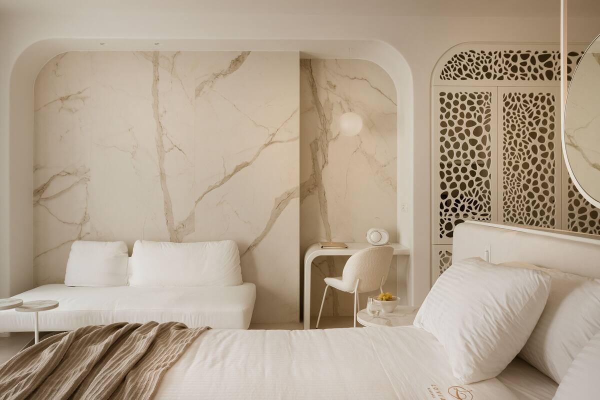 A modern bedroom with a marble wall, a bed with white bedding, a bedside table with a lamp, and a decorative screen.