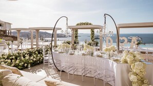 Decoración para boda al aire libre con vista al mar, que incluye una mesa blanca con sillas transparentes, arreglos florales y una estructura de toldo.