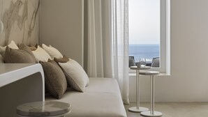 Un dormitorio moderno con sofá, almohadas, una pequeña mesa redonda y vistas al océano a través de cortinas translúcidas.