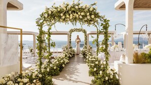 Un pasillo de bodas con arcos florales blancos y sillas, que conduce a vistas del mar.