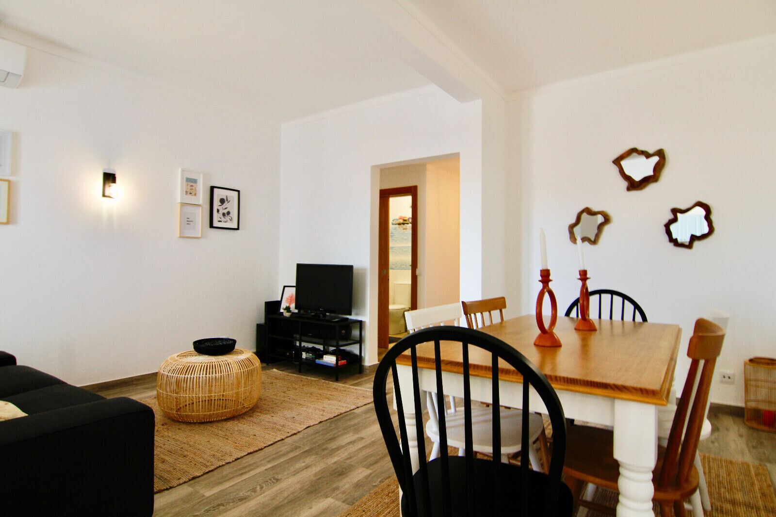 A living room with a black sofa, a TV on a black stand, a dining table with chairs, and a wicker ottoman.
