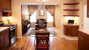 A modern kitchen with wooden cabinets, a dining area with a glass table, and a living room with a sofa and curtains.