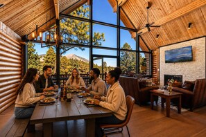 Un groupe de personnes est assis à une table en bois, devant de grandes fenêtres qui offrent une vue sur la montagne, et prend son repas.