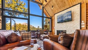 A spacious living room with leather sofas, a coffee table with glasses, and a large window offering a mountain view. - Renovated | Famous Cabin | Hot Tub | AC | Mtn View (Flagstaff)