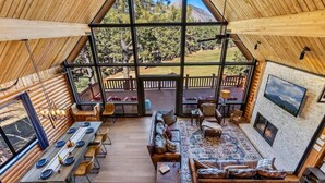 A spacious interior with a long dining table, comfortable seating, a fireplace, and a large window offering a view of the outdoors. - Renovated | Famous Cabin | Hot Tub | AC | Mtn View (Flagstaff)