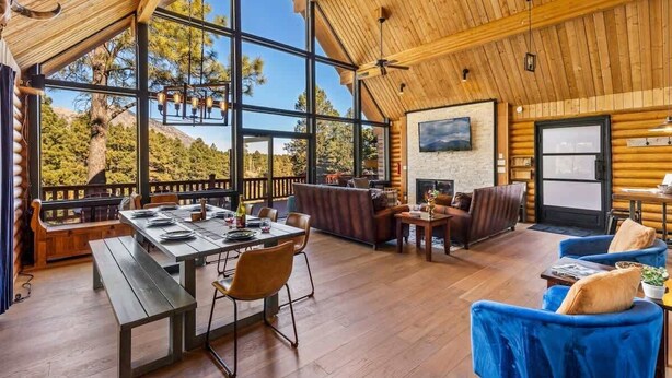 A spacious interior with a wooden ceiling, large windows offering a view of trees, a dining area with a table and chairs, and a seating area with sofas and a fireplace. - Renovated | Famous Cabin | Hot Tub | AC | Mtn View (Flagstaff)