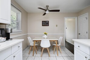 A modern kitchen with white cabinetry, a dining table with four chairs, a ceiling fan, and a window with blinds.