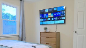 Une chambre à coucher comprenant un lit, un téléviseur fixé au mur qui diffuse différentes chaînes, une commode en bois et une fenêtre donnant sur des arbres.