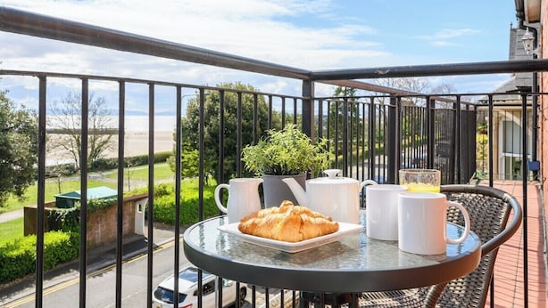 A balcony with a table set for breakfast, including a croissant, two white mugs, and a pitcher of orange juice.