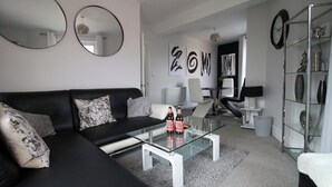 A modern living room with a black leather sofa, a glass coffee table, and a white side table. There are two round mirrors on the wall, a bookshelf, and a radiator.