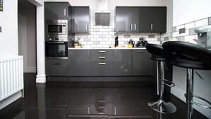 A modern kitchen with dark grey cabinets, a stainless steel oven, a microwave, and a coffee machine. There are two black bar stools with chrome legs.