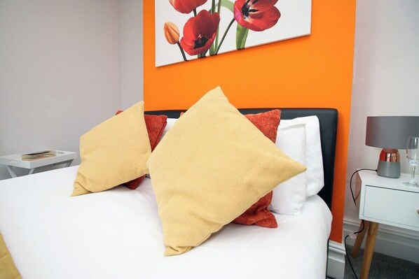 Una cama con almohadas blancas y amarillas, una pared naranja y un cuadro con flores rojas.