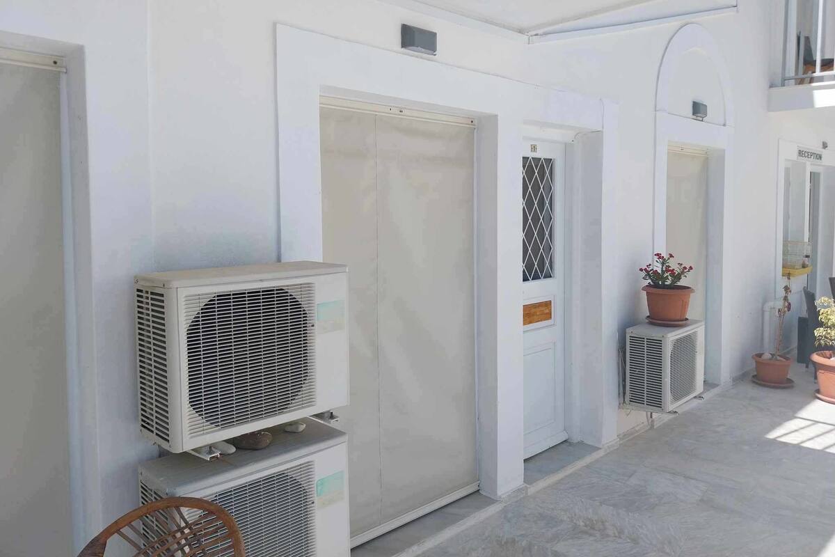 Un pasillo con paredes blancas, puertas y unidades de aire acondicionado. Hay plantas en macetas y una silla de mimbre.