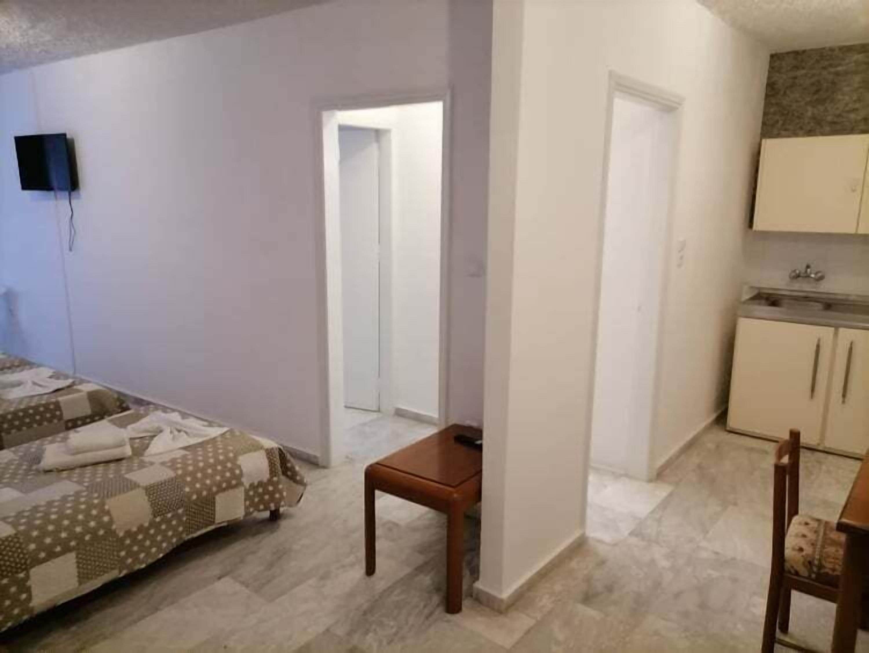 A bedroom with a bed, a small table, and a kitchenette area with cabinets and a sink.