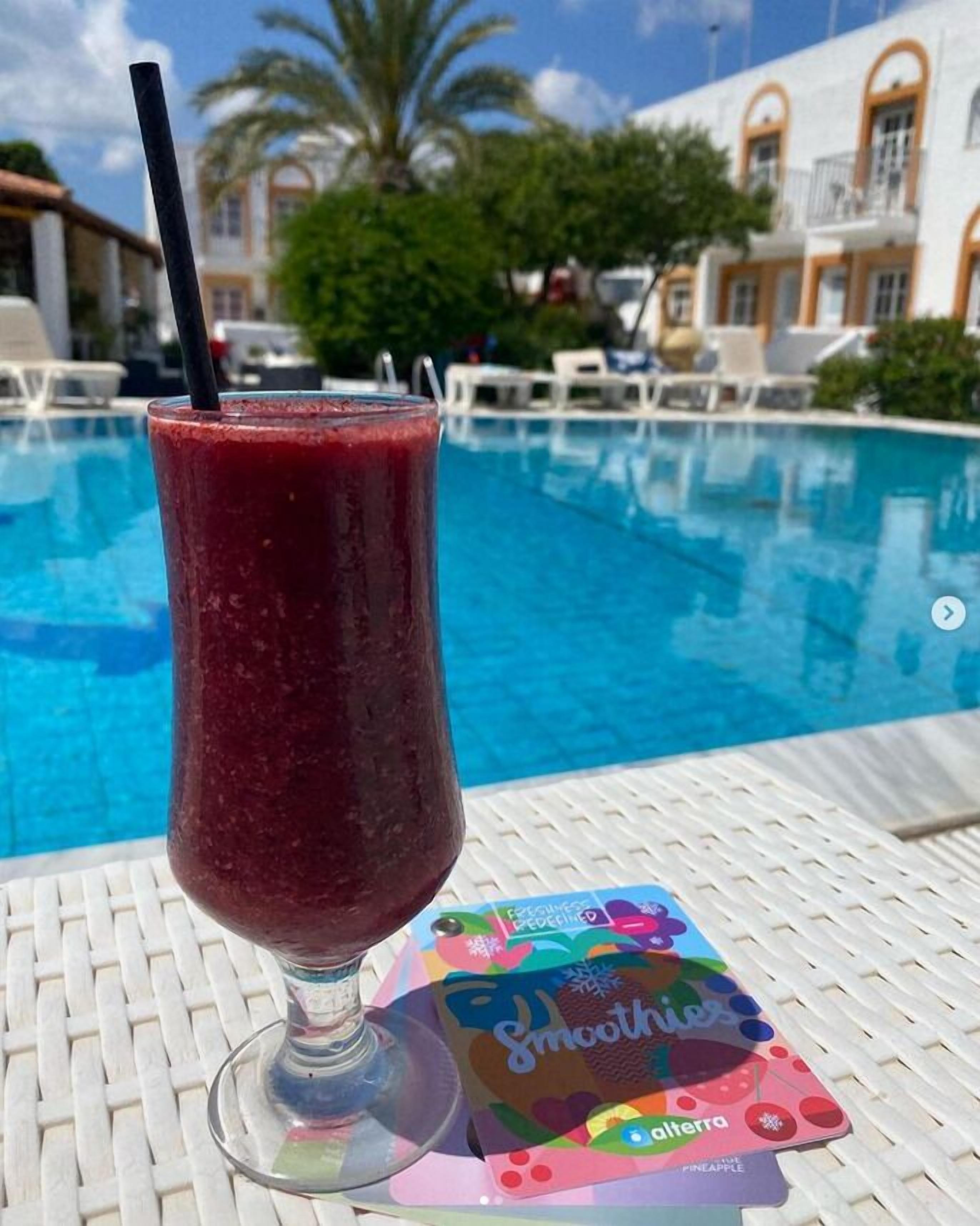 A glass of red smoothie with a straw on a white table by a pool. A colorful brochure titled 'Smoothies' is on the table.