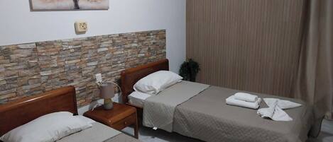 A hotel room with two beds, a stone wall, a painting, a bedside table with a lamp, and a window with curtains.