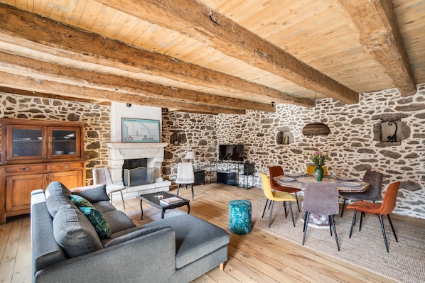 A rustic interior with a stone wall, wooden beams, a fireplace, a sofa, a dining table with chairs, and a cabinet.