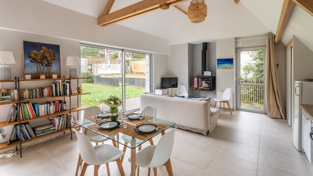 A spacious living area with a glass dining table, white chairs, and a bookshelf filled with books. A kitchen area with a stove and a dining table is visible in the background. - Ar Chadenn - Maison d'architecte (Saint-Cast-le-Guildo)