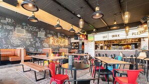 L’intérieur d’un restaurant présente des tables en bois, des chaises colorées et un mur noir où sont dessinés des motifs liés à la nourriture.