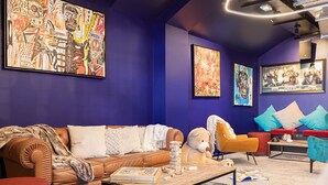 Un salon moderne avec un canapé en cuir marron, un fauteuil jaune et un pouf rouge. Les murs sont ornés de tableaux abstraits et une pile de livres est posée sur la table basse.