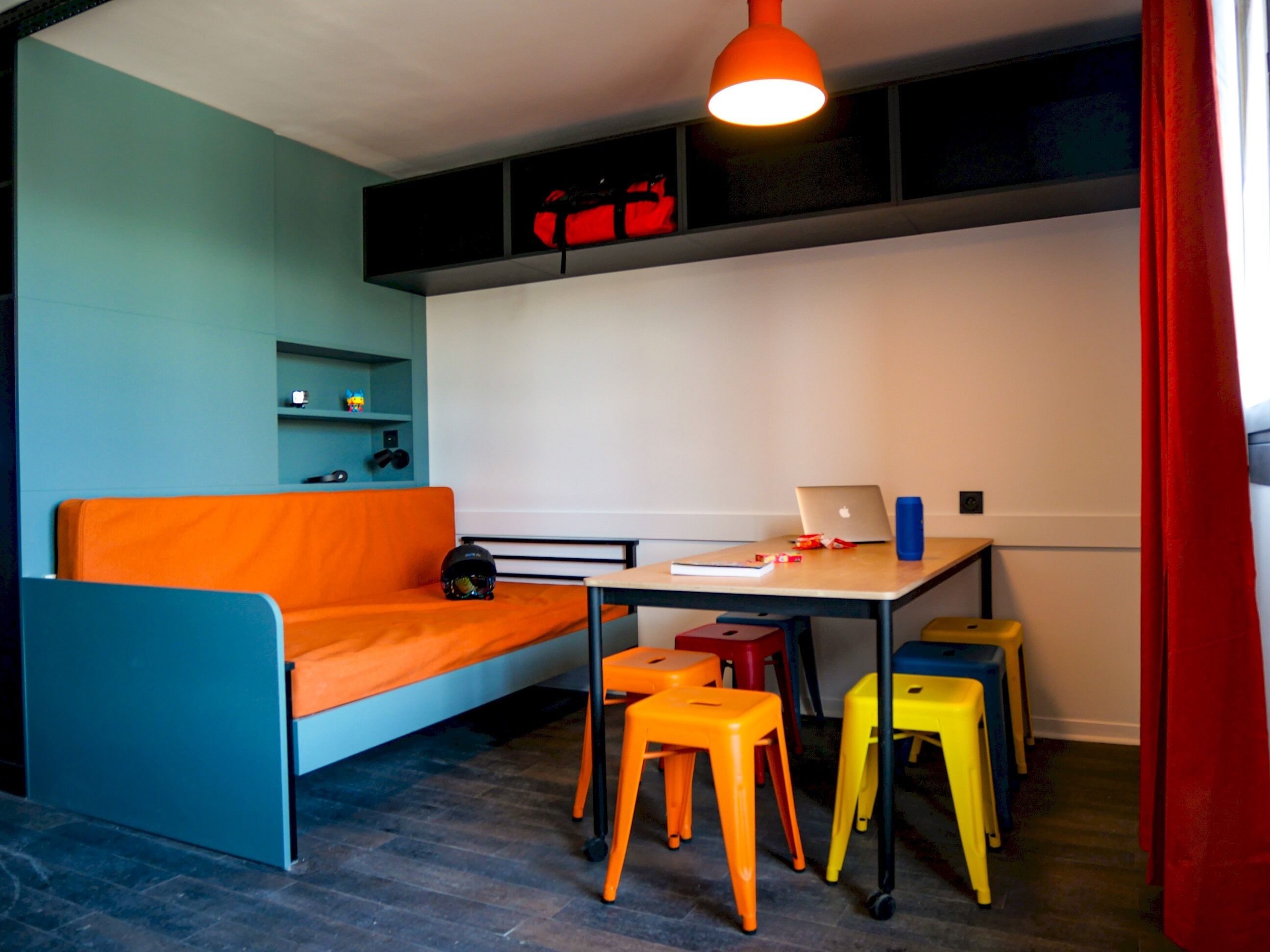 A room with a blue sofa, a desk with a laptop, colorful stools, and a red curtain.