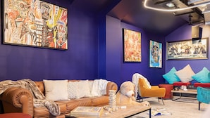 Ein modernes Wohnzimmer mit einem braunen Ledersofa, einem gelben Sessel und einem roten Hocker. An den Wänden hängen abstrakte Gemälde und auf dem Couchtisch liegt ein Stapel Bücher.