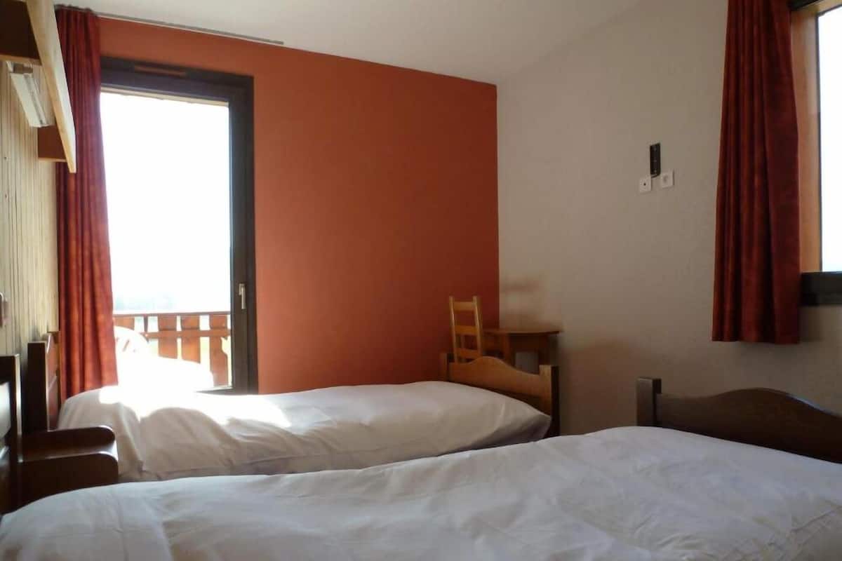A room with two beds, a wooden chair, a window with curtains, and a balcony.