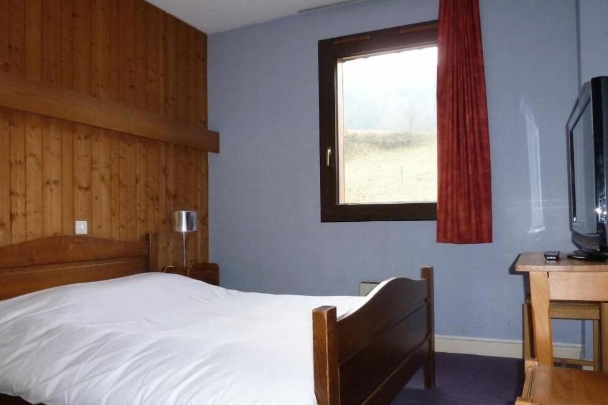 A bedroom with a wooden headboard, a single bed, a small table with a television, and a window with a red curtain.