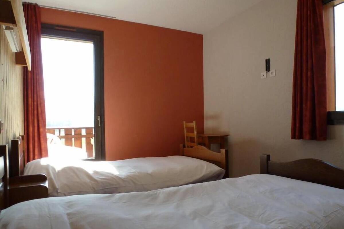 A room with two beds, a wooden chair, a window with curtains, and a balcony visible through the window.