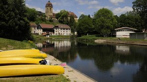 A calm river with yellow canoes lined up on the bank, a historic building with a tower in the background, and lush greenery surrounding the area. - Espace Morteau (Morteau)