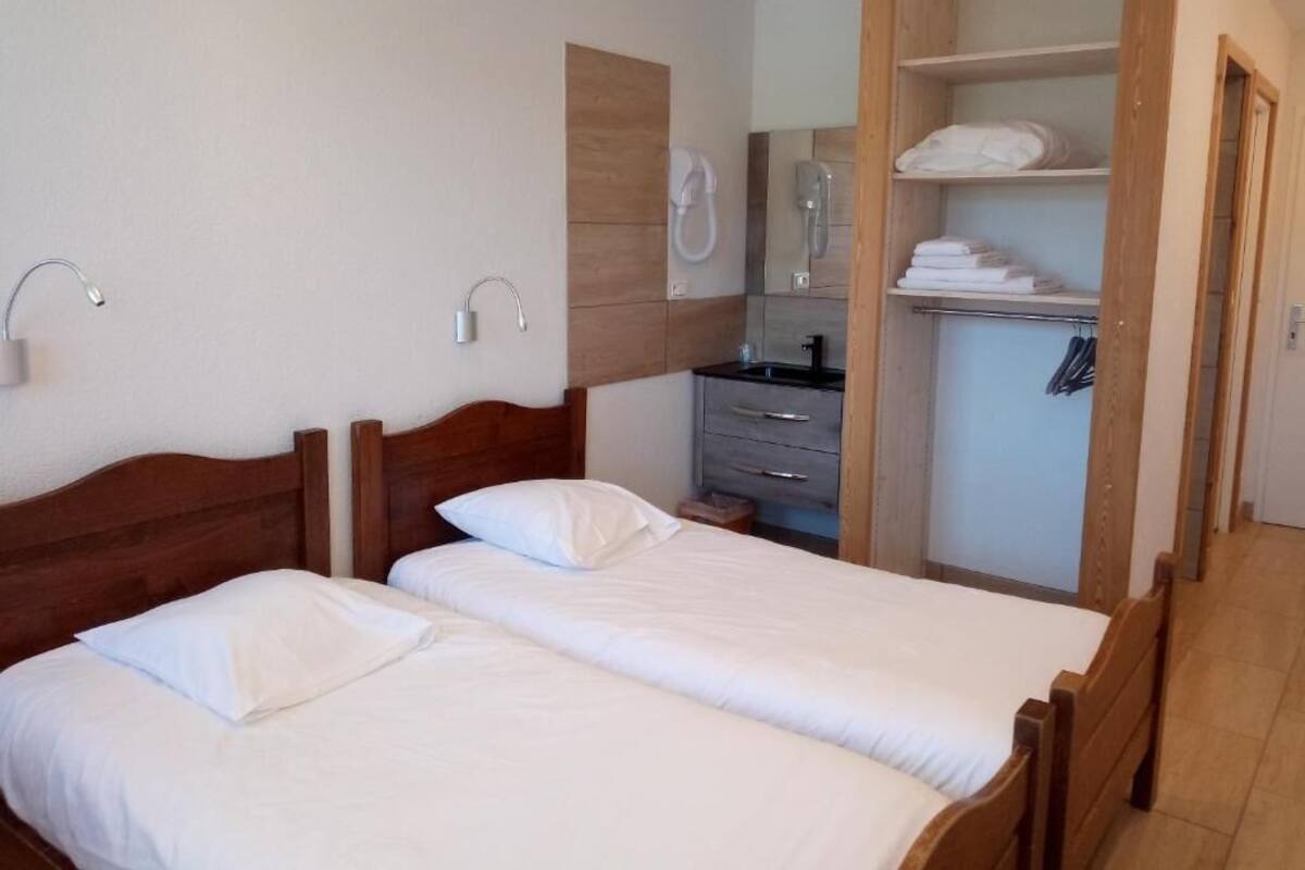 A bedroom with two beds, a wooden wardrobe, a mirror, and a bathroom with a sink and towel rack.