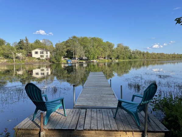 A wooden dock with two chairs facing a calm lake, surrounded by trees and a house in the background.