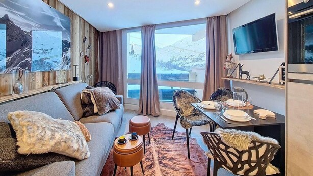 Una sala de estar moderna con sofá, mesa de comedor y sillas, un ventanal con cortinas y un televisor de pantalla plana montado en la pared.