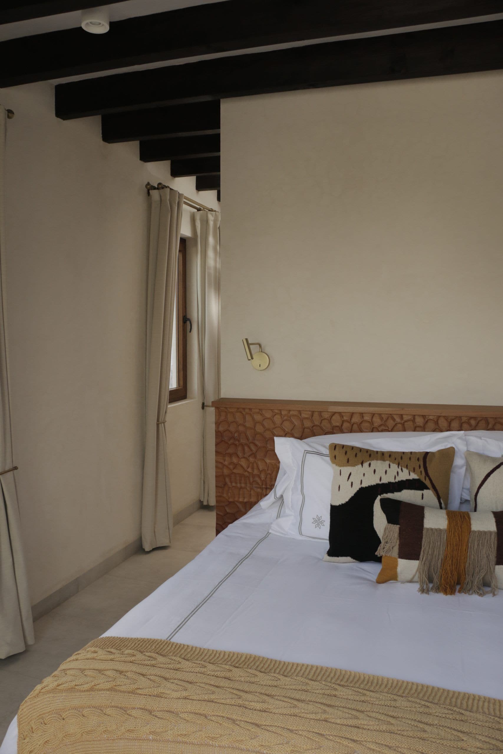 A hotel room with a bed, a headboard, a window with curtains, and a wall-mounted light fixture.