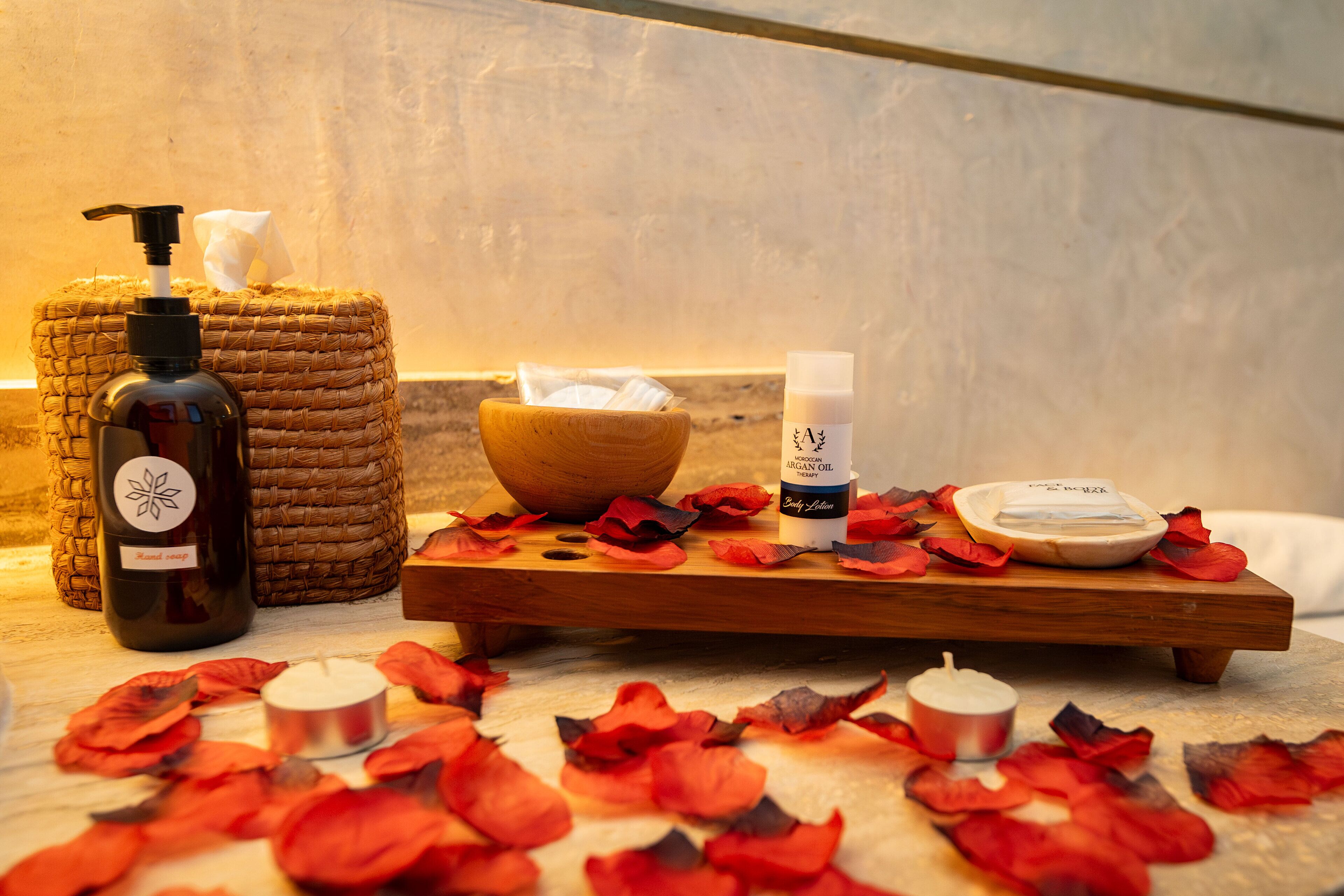 A wooden tray with a bottle of argan oil, a bowl, and a candle on a marble surface surrounded by rose petals.
