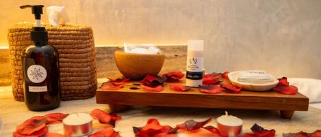 A wooden tray with a bottle of argan oil, a bowl, and a candle on a marble surface surrounded by rose petals.